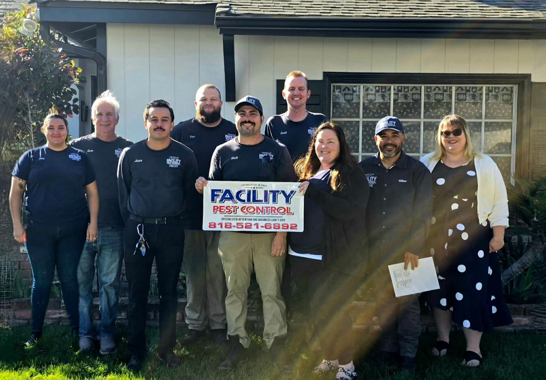 The nine person Facility Pest Control team in navy uniforms in front of a Reseda home, holding the company sign
